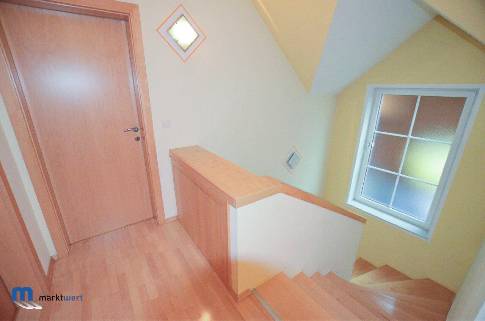 Heller Treppenabsatz mit Holzgeländer und Fenster, das Tageslicht hereinlässt.