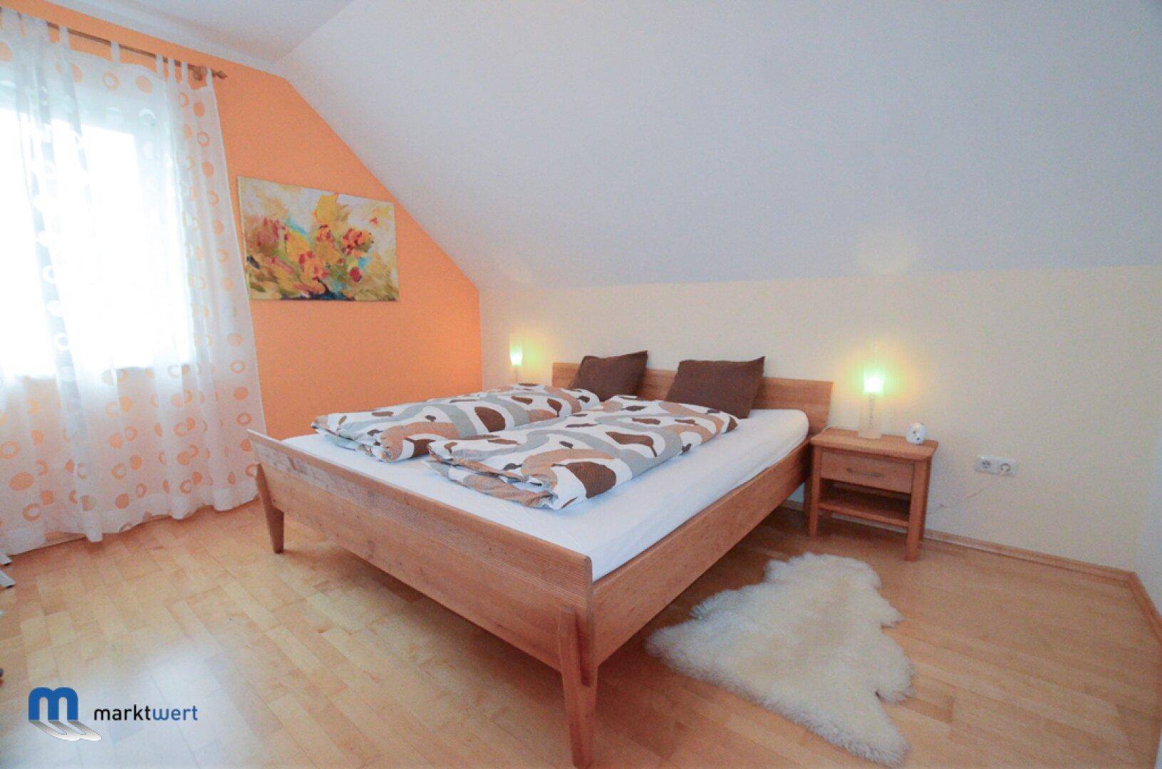 Helles Schlafzimmer mit Holzbett, Fenster, orangefarbener Akzentwand und Dachschrägen.