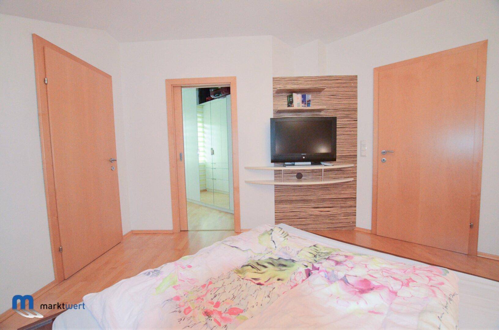 Schlafzimmer mit Doppelbett, Fernseher an der Wand und Zugang zum begehbaren Kleiderschrank.