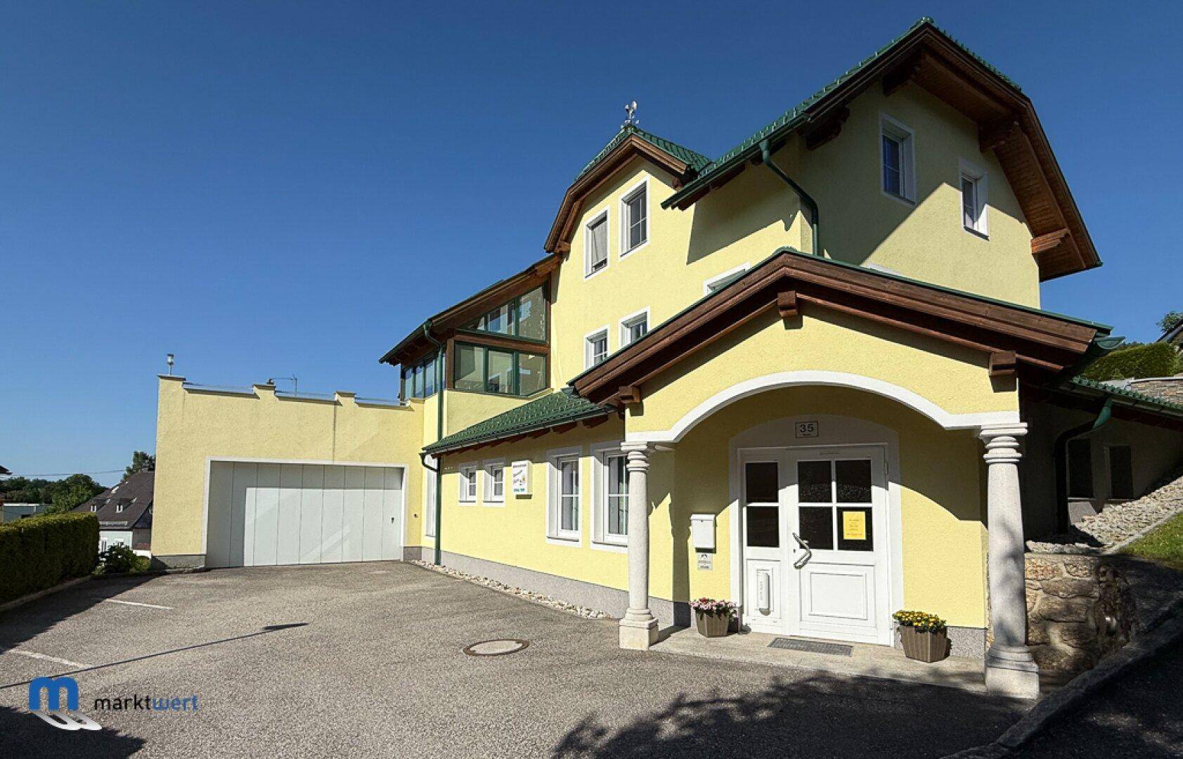 Ansicht des Hauses mit gelber Fassade, grünem Dach und Säulen am Eingangsbereich.