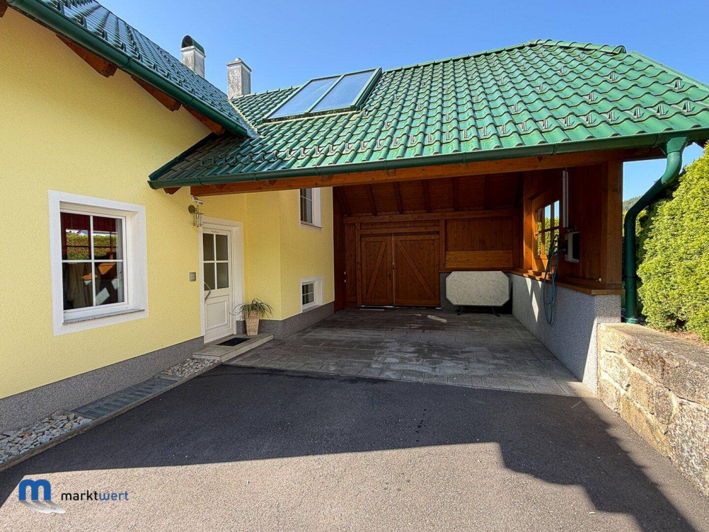 Überdachter Stellplatz mit Holzbalken und Zugang zum Haus, umgeben von grüner Hecke.