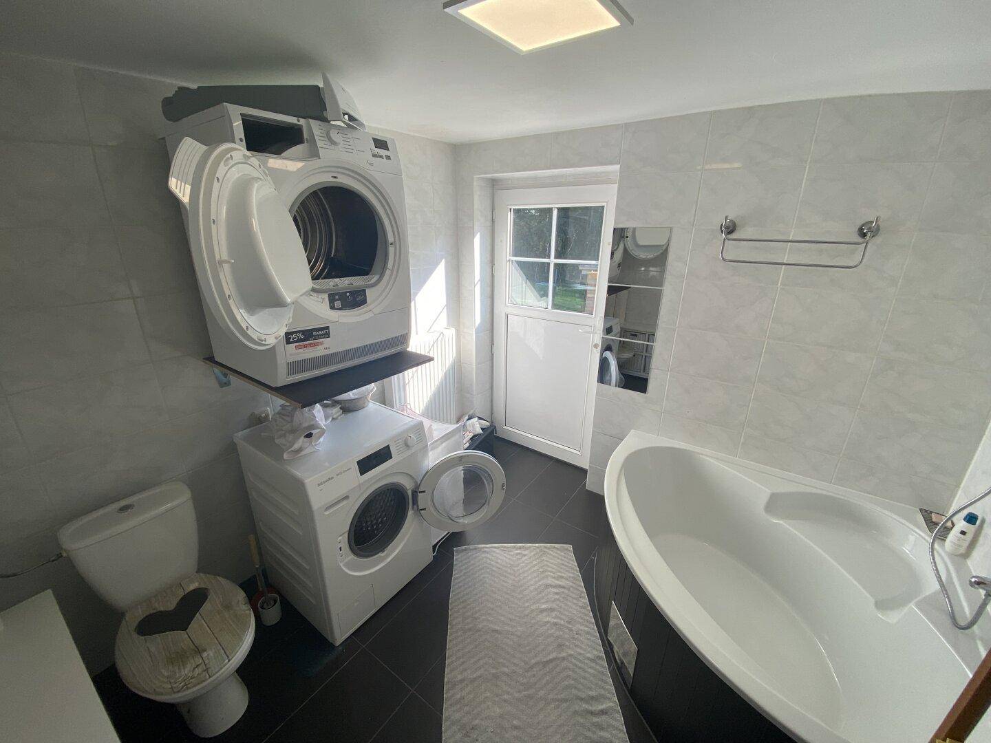 Helles Badezimmer mit Eckbadewanne, WC und gestapelter Waschmaschine und Trockner.