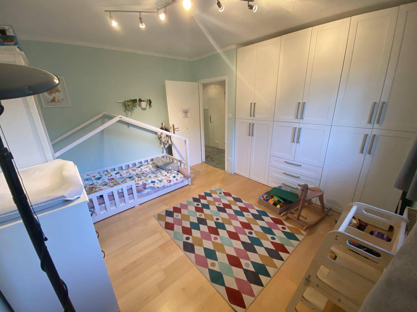 Freundliches Kinderzimmer mit hellem Holzboden, weißem Kleiderschrank und Spielteppich.