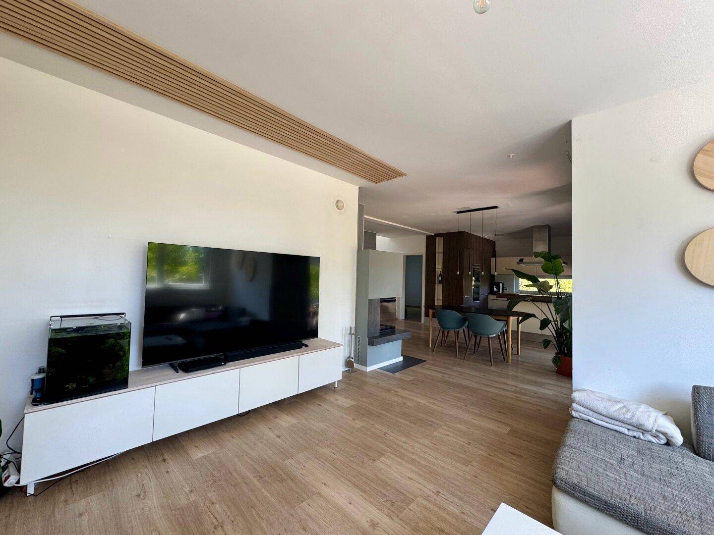 Geräumiges Wohnzimmer mit großem Fernseher, Holzboden und offenem Übergang zum Essbereich.