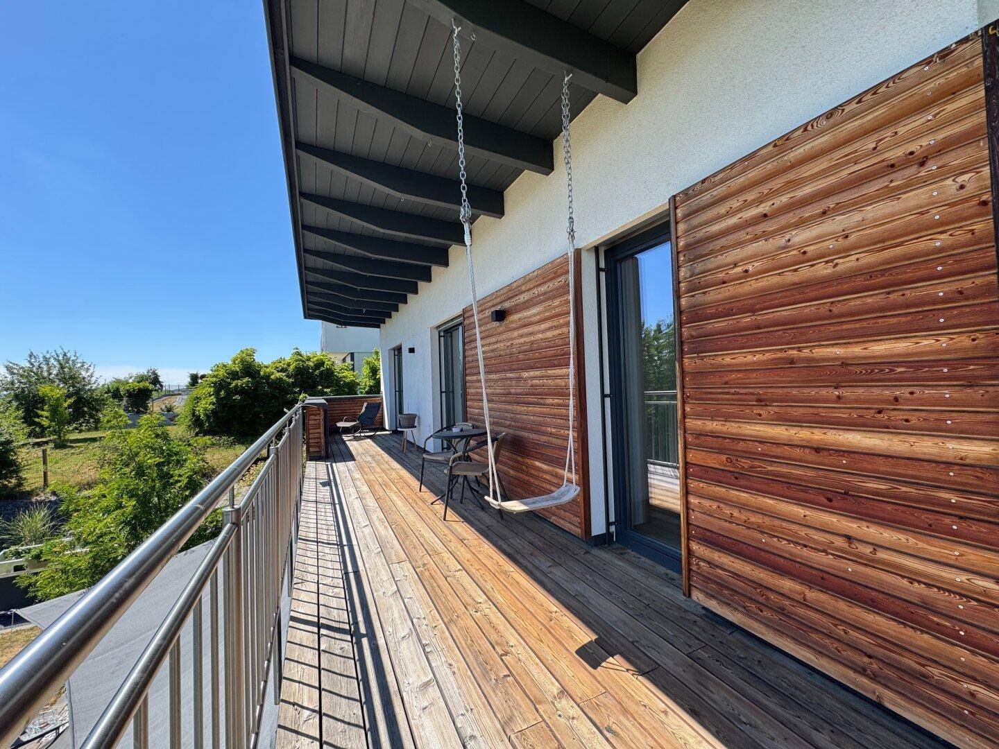 Heller Balkon mit Holzboden, Schaukel und Sitzgelegenheiten, direkt am Haus mit Holzfassade.