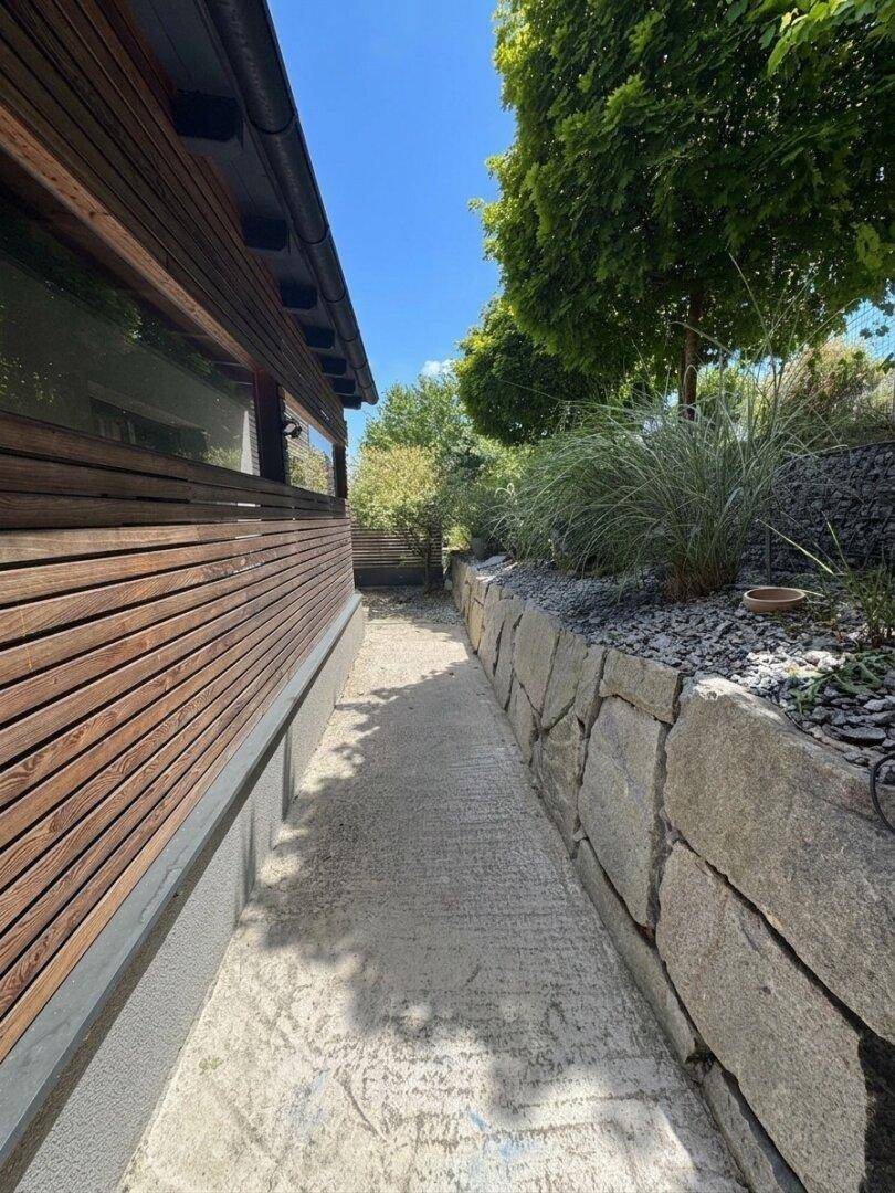 Schmaler Betonweg entlang des Hauses mit Natursteinmauer und Holzfassade.