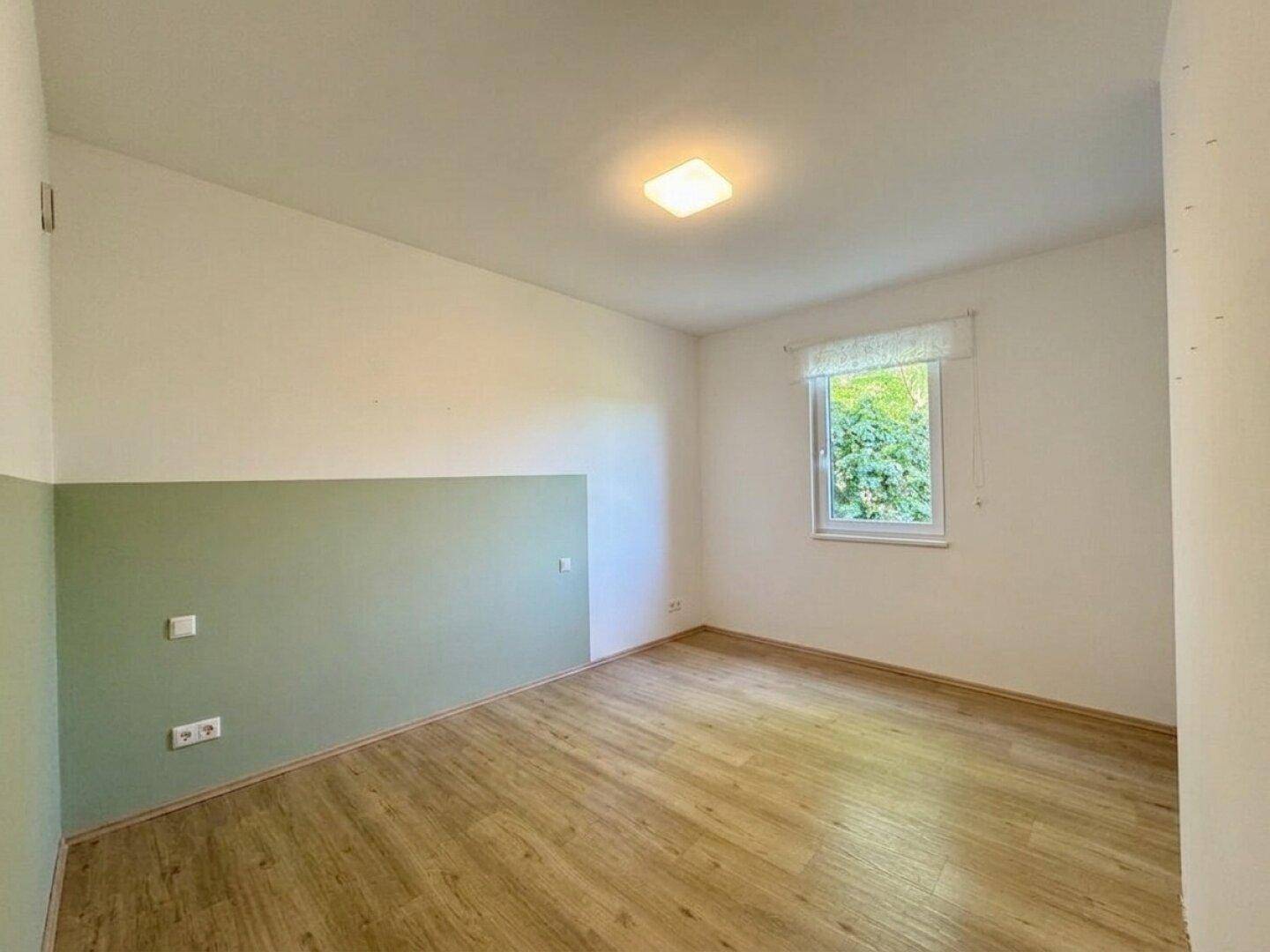Helles Schlafzimmer mit Holzboden, grüner Wandgestaltung und Fenster mit Blick ins Grüne.