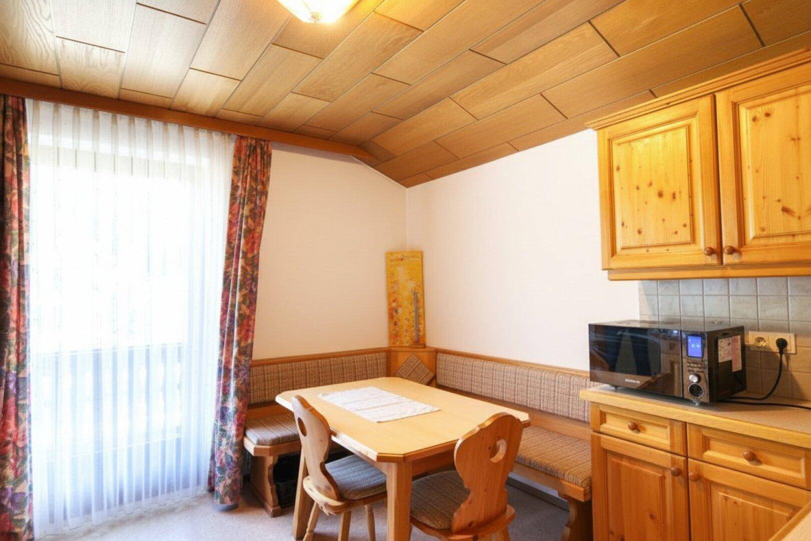 Traditionelle Wohnküche mit Holzeinbauten, Eckbank, Esstisch und Mikrowelle am Fenster.