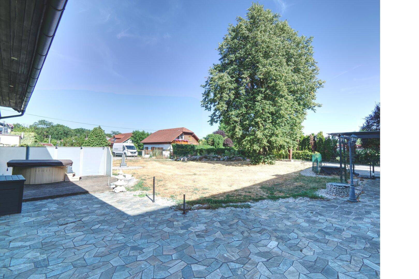 Gepflasterte Terrasse mit Whirlpool und großem Baum im Gartenbereich.