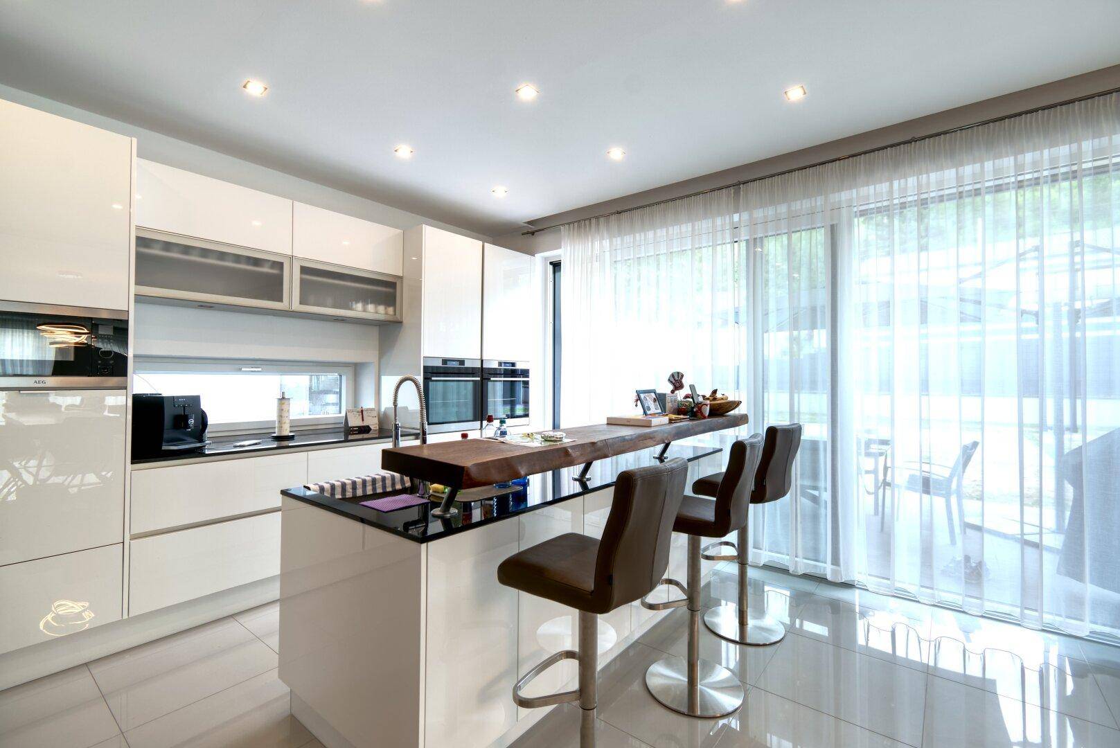 Moderne Küche mit weißen Hochglanzfronten, Kücheninsel und direktem Zugang zur Terrasse durch große Fenster.