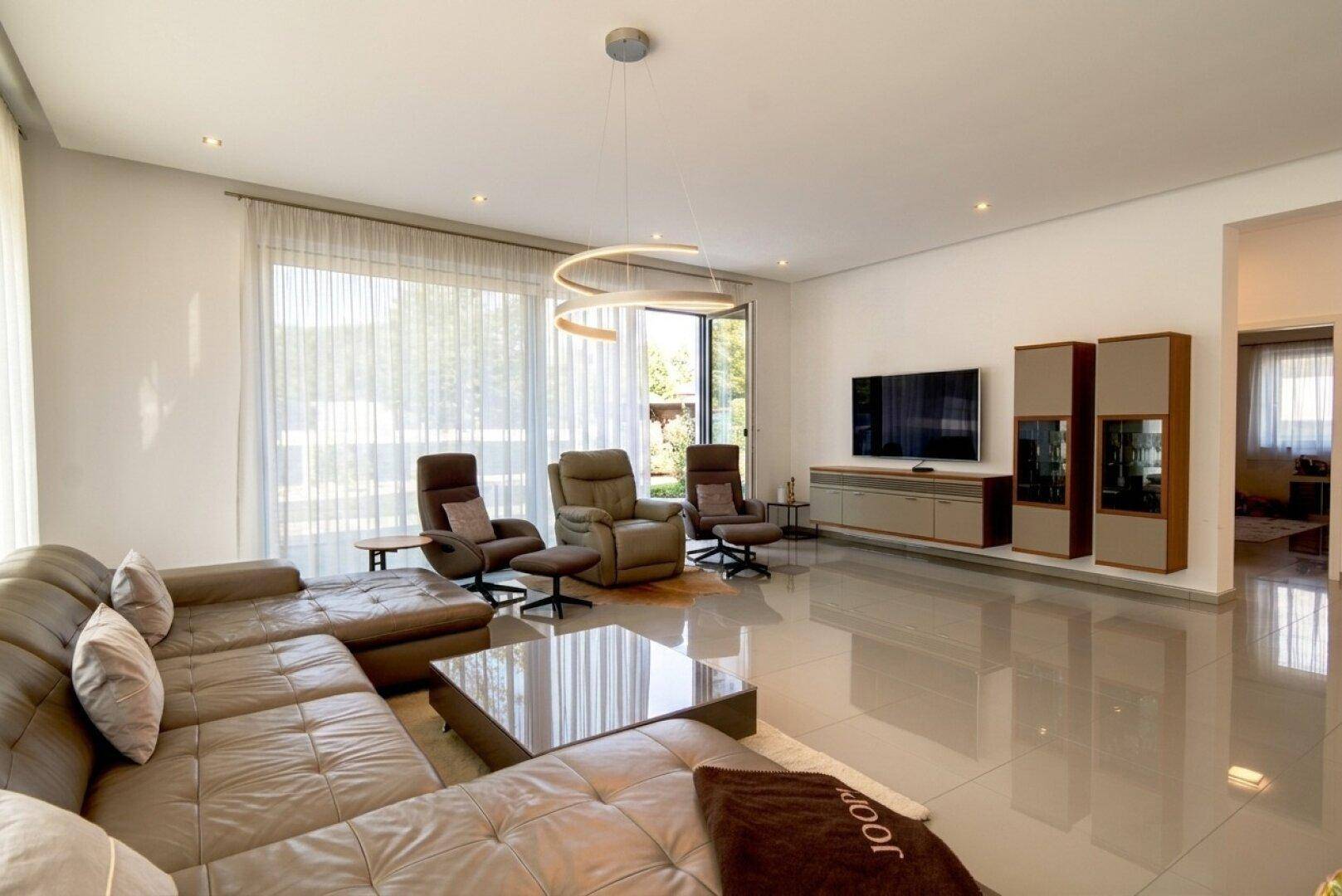 Geräumiges Wohnzimmer mit bequemer Sofalandschaft, Fernseher und großen Fenstern für viel Tageslicht.
