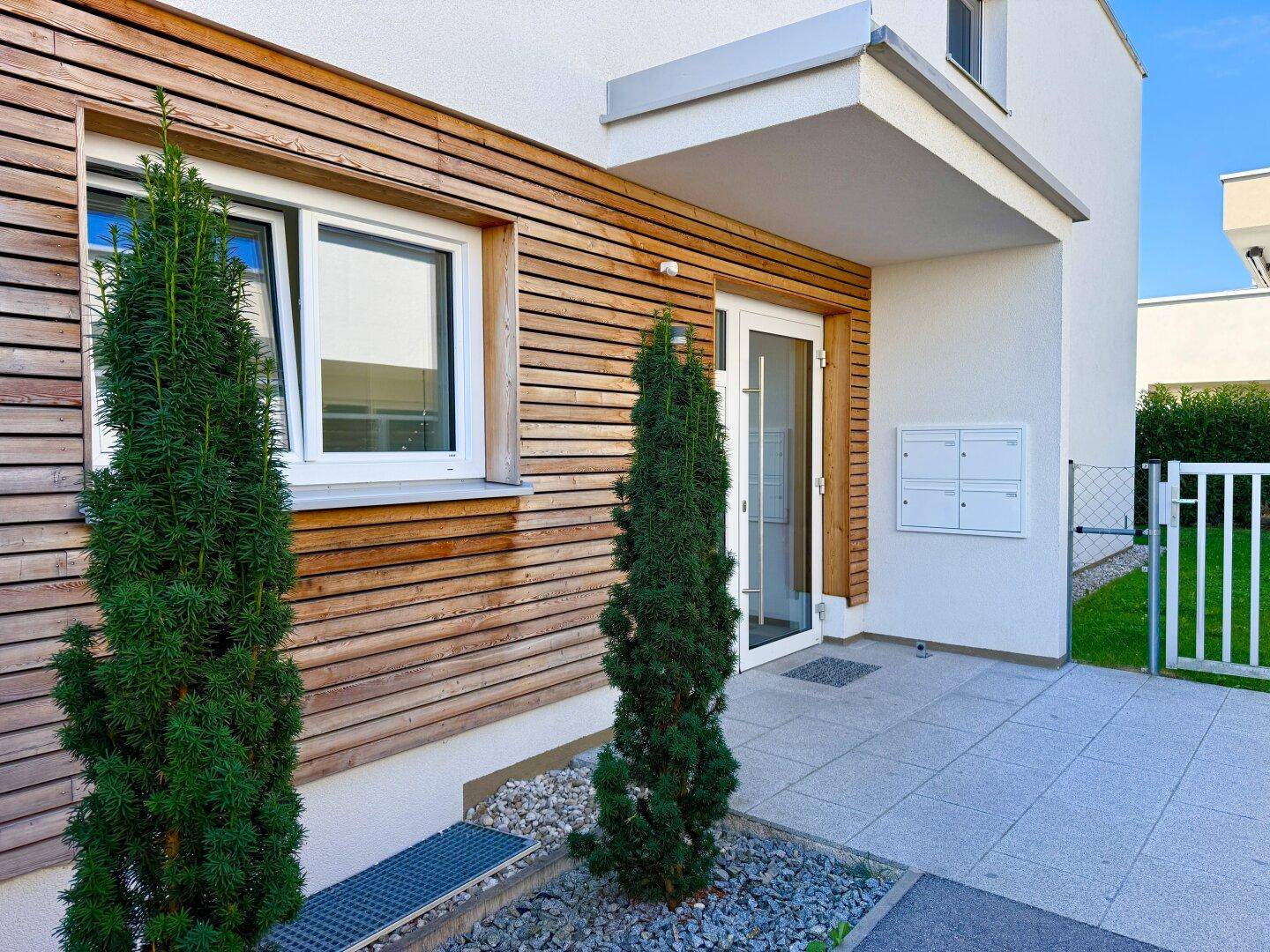 Moderner Hauseingang mit Holzfassade, großen Fenstern und gepflegter Bepflanzung.