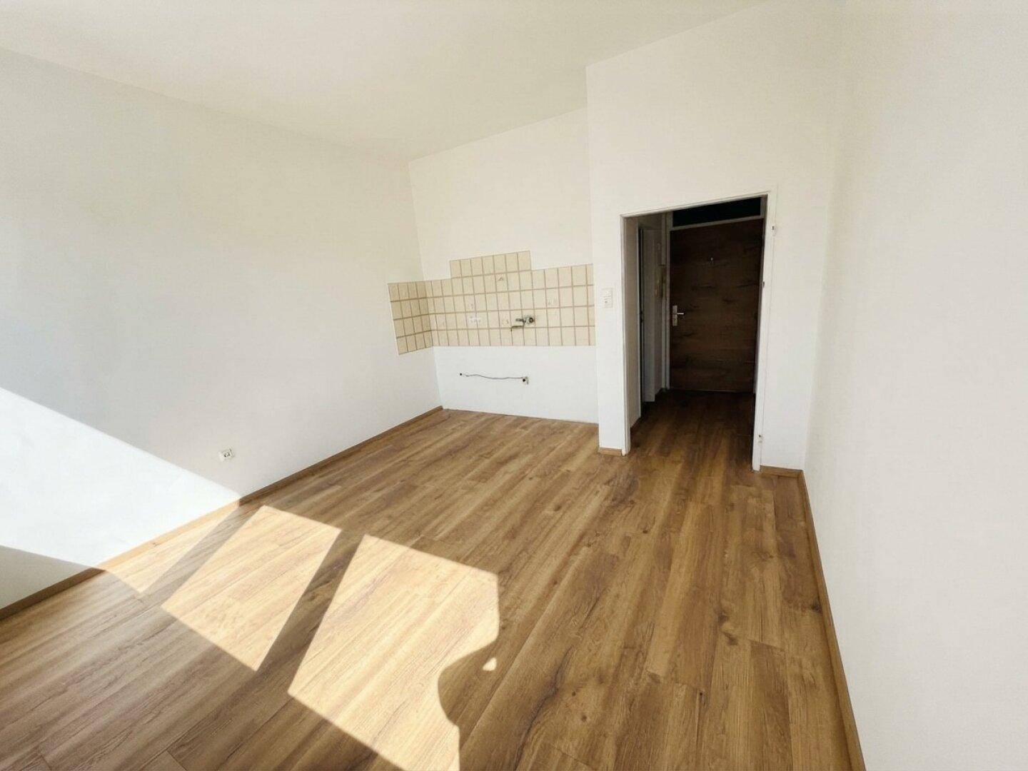 Leerer Küchenbereich mit Laminatboden, weißen Wänden und Fliesen als Spritzschutz.