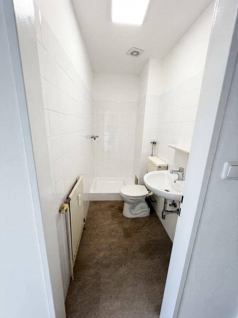 Schmales Badezimmer mit weißen Fliesen, Dusche, WC, Waschbecken und Heizkörper.