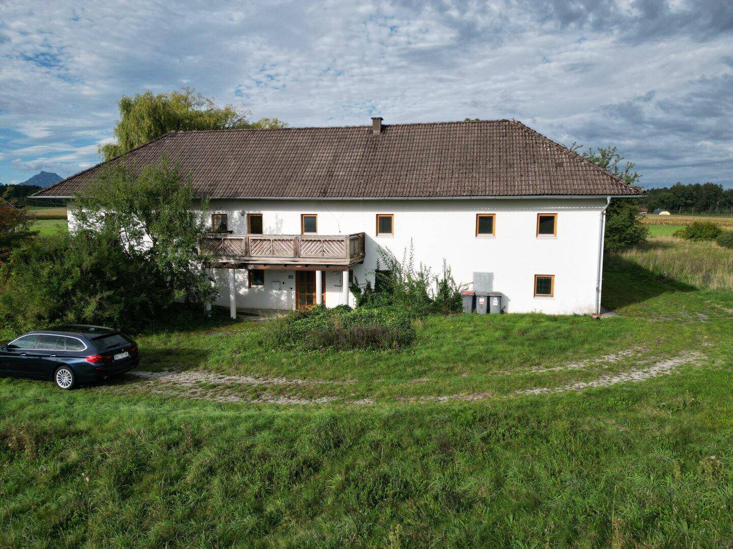 Fassade des Landhauses mit weißem Putz, Holzbalkon und geparktem Fahrzeug auf dem Rasen.