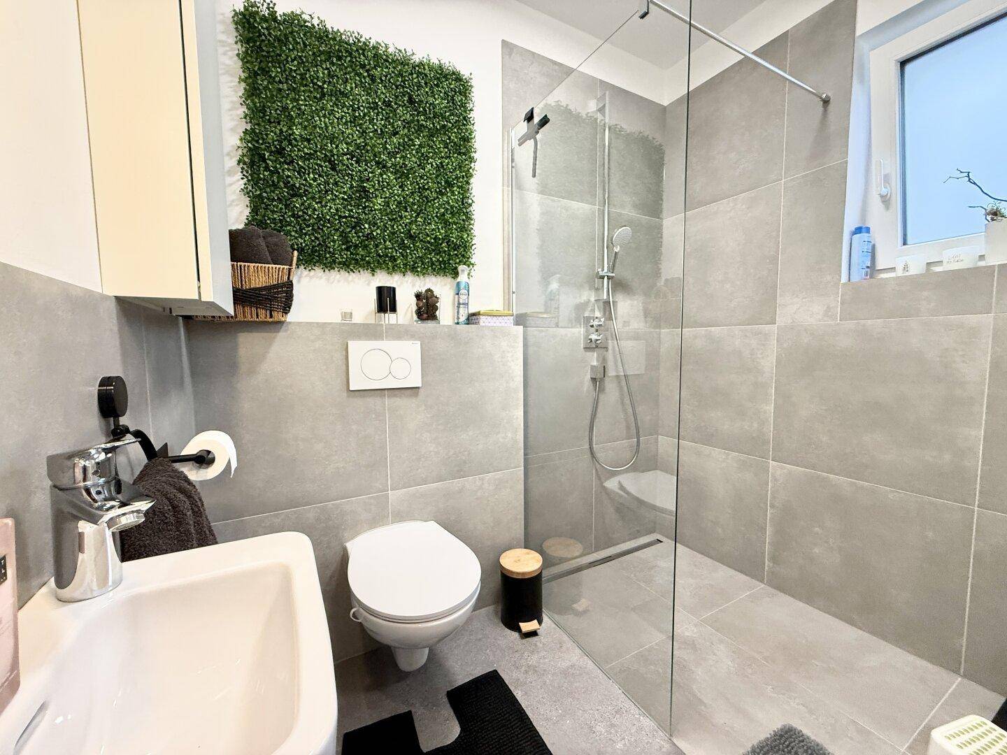 Modernes Badezimmer mit großformatigen grauen Fliesen, wandhängendem WC und begehbarer Dusche.