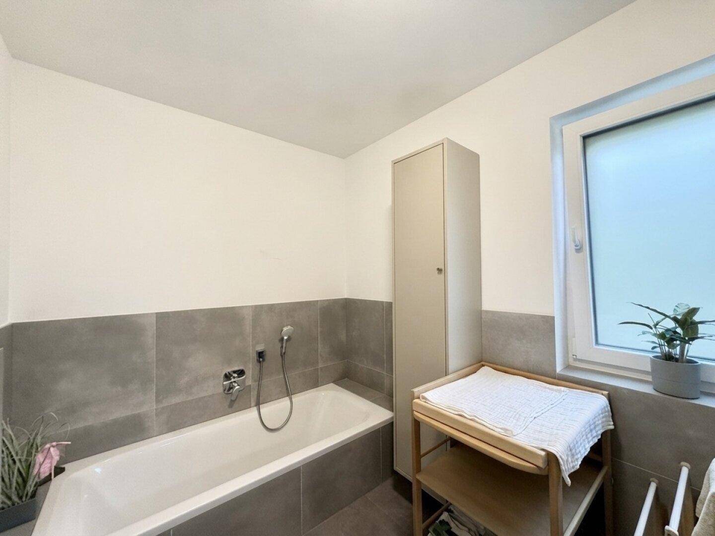 Helles Badezimmer mit Badewanne, Fenster und schlichten grauen Wandfliesen.