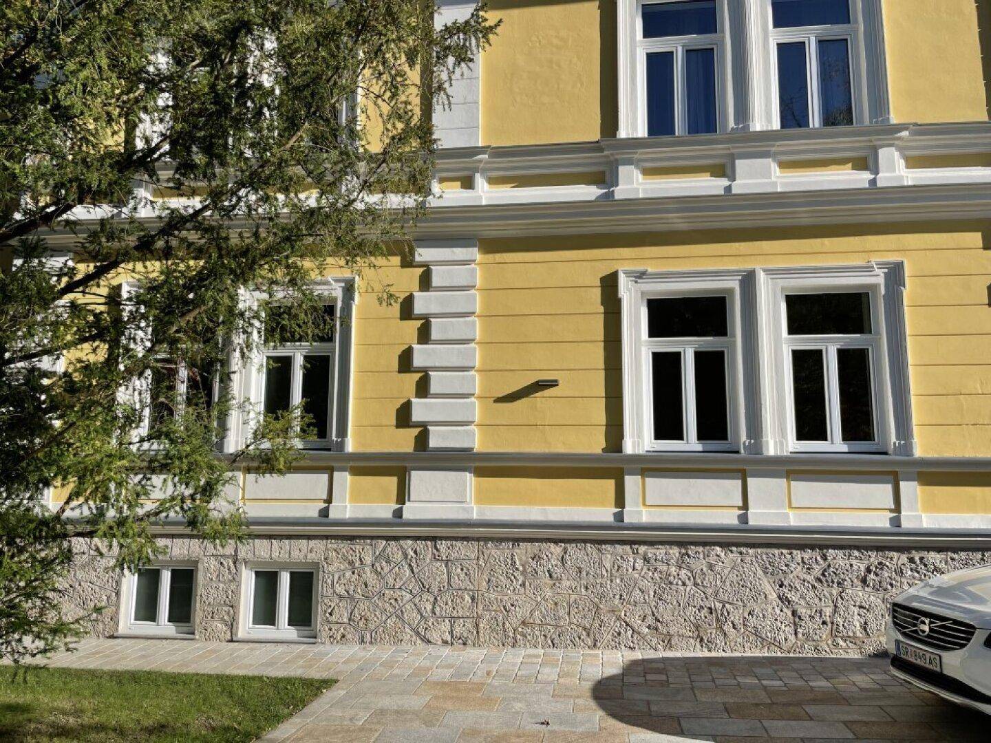Nahaufnahme der gelben Fassade mit weißen Fensterrahmen, dekorativen Stuckelementen und einem gepflasterten Bereich.