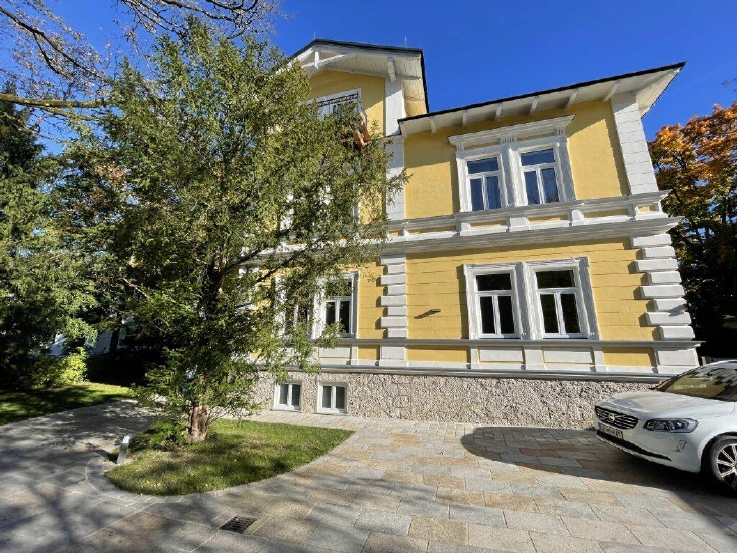 Seitenansicht der historischen gelben Villa mit gepflegtem Vorgarten, einer Einfahrt und einem geparkten Fahrzeug.