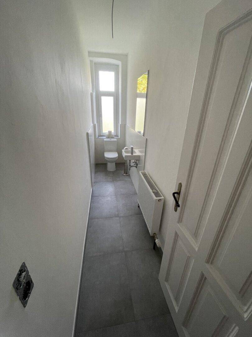 Schmaler Toilettenraum mit grauem Fliesenboden, einem Fenster und einem Heizkörper unter dem Fenster.