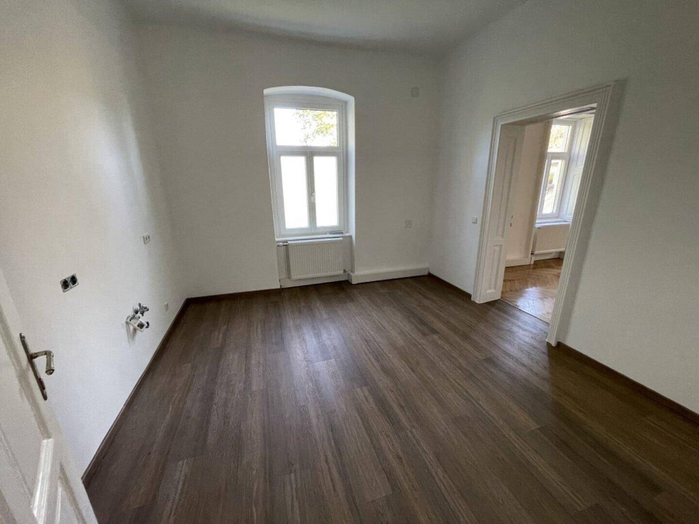 Zimmer mit dunklem Holzboden, einem Fenster und vorbereiteten Anschlüssen für eine mögliche Küchenzeile.