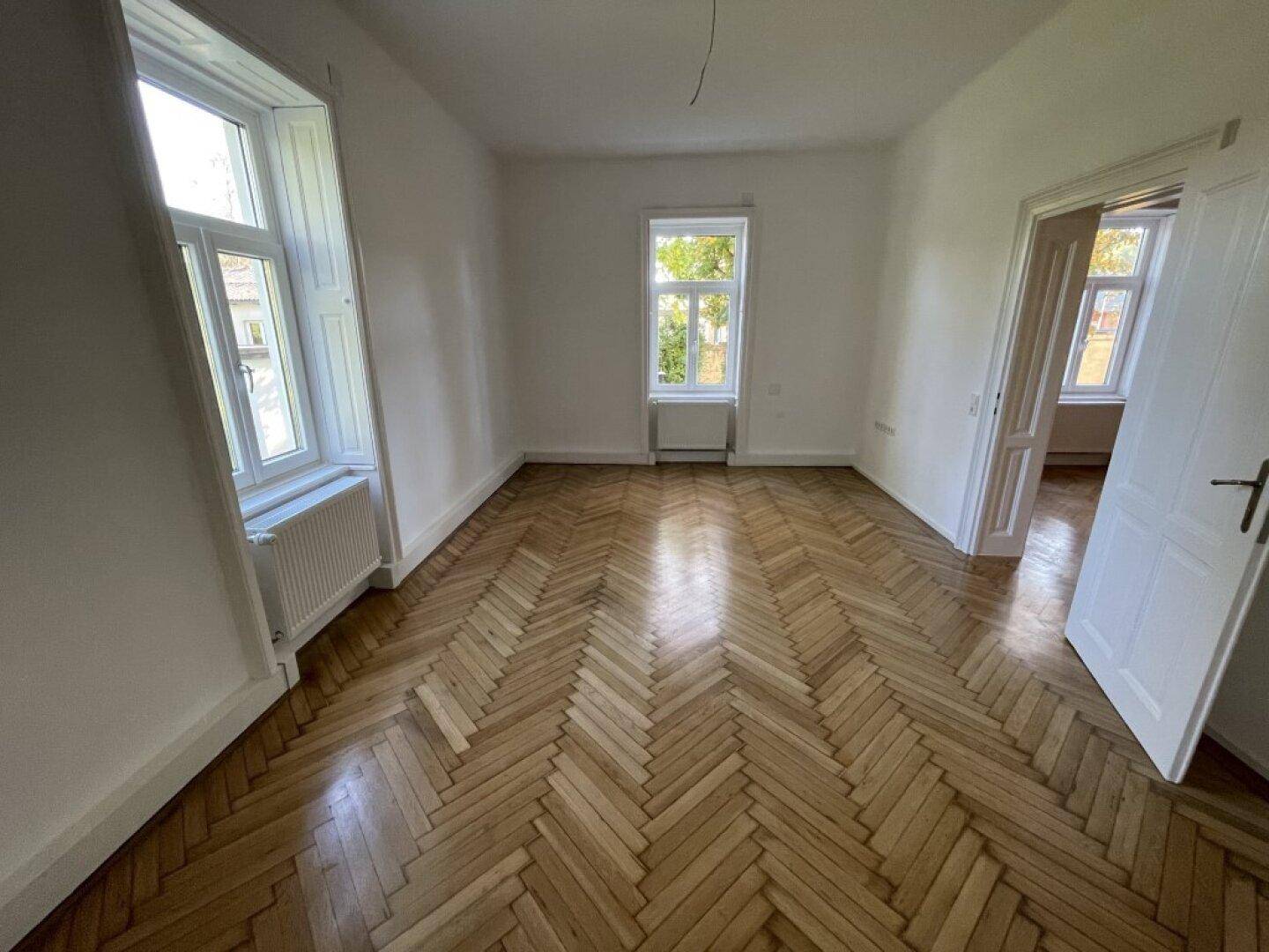 Heller Raum mit Fischgrätparkett, hohen Fenstern und Blick ins Grüne, bereit zur individuellen Gestaltung.