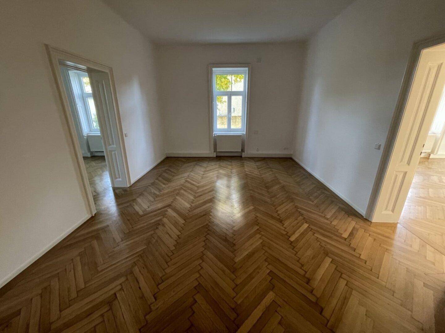 Geräumiges Zimmer mit klassischem Fischgrätparkett und einem großen Fenster, das viel Tageslicht hereinlässt.