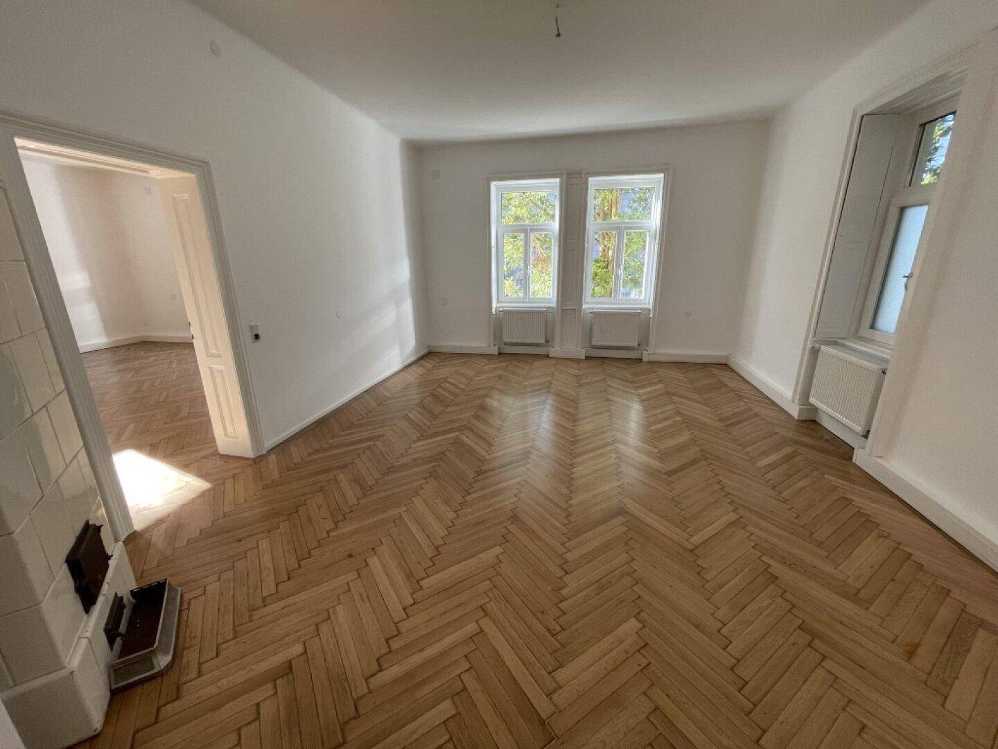 Helles Zimmer mit Fischgrätparkett, zwei Fenstern und einem traditionellen Kachelofen als Blickfang.