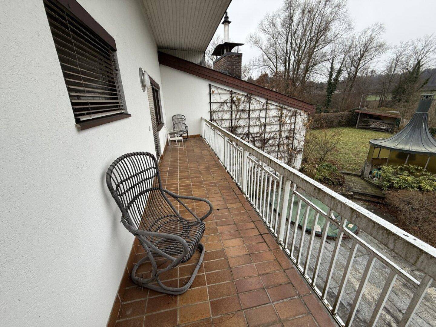 Balkon mit Terrakotta-Fliesen und Metallgeländer, ausgestattet mit einem Korbstuhl und Blick ins Grüne.