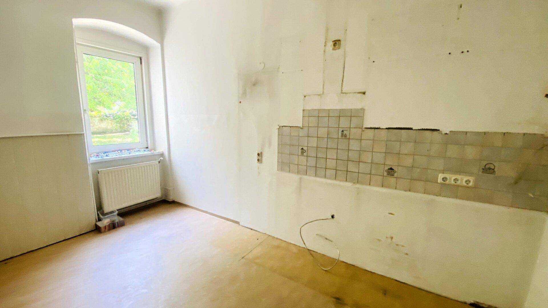 Leere Küche mit alten Fliesen an der Wand, Heizkörper und Fenster mit Blick ins Grüne.