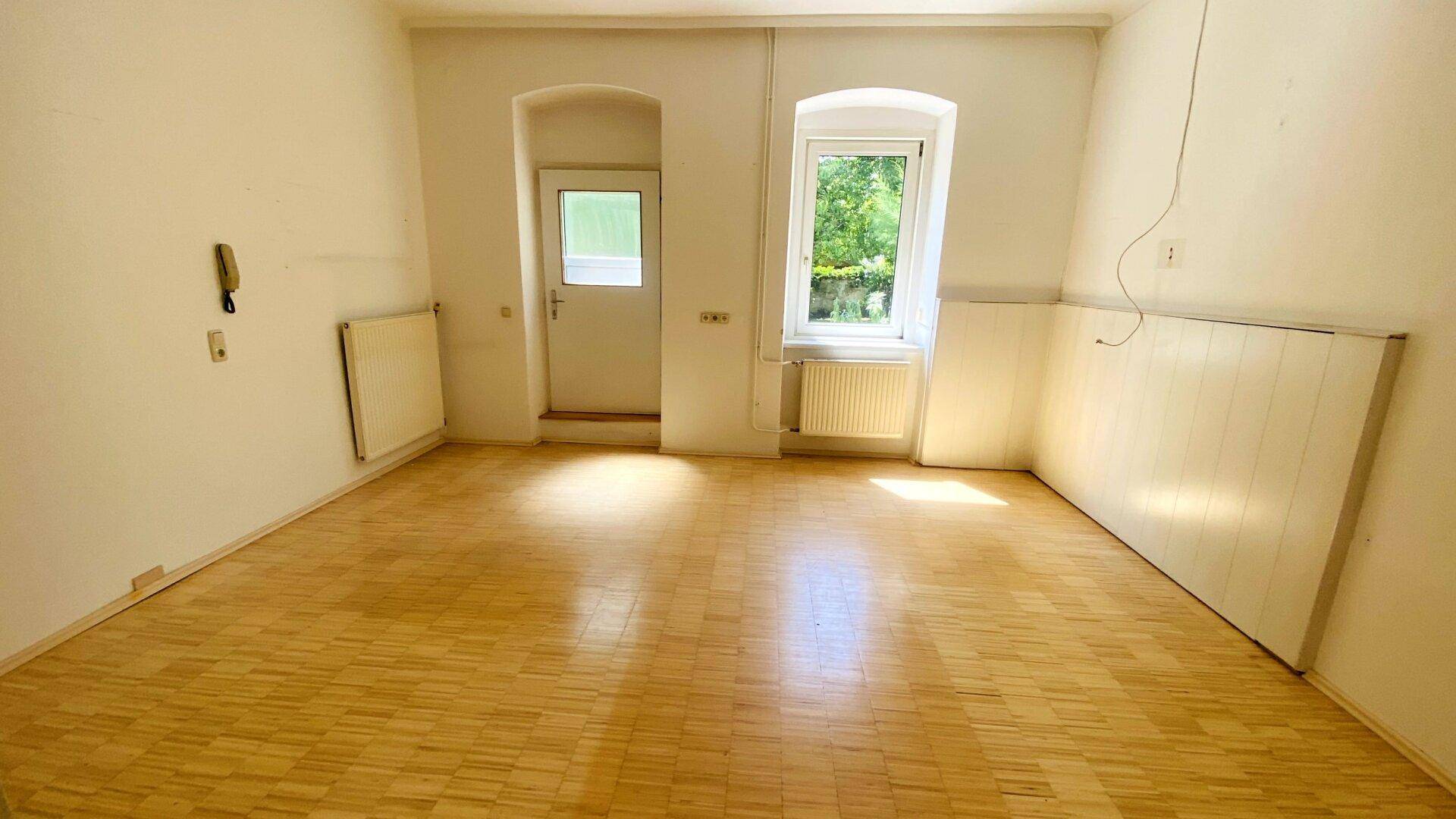 Helles Wohnzimmer mit Parkettboden, Heizkörpern und Blick ins Grüne durch das Fenster.