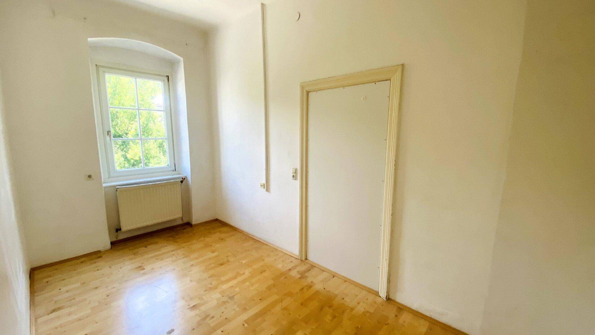 Helles Schlafzimmer mit Parkettboden, weißer Tür und Fenster mit Blick ins Grüne.