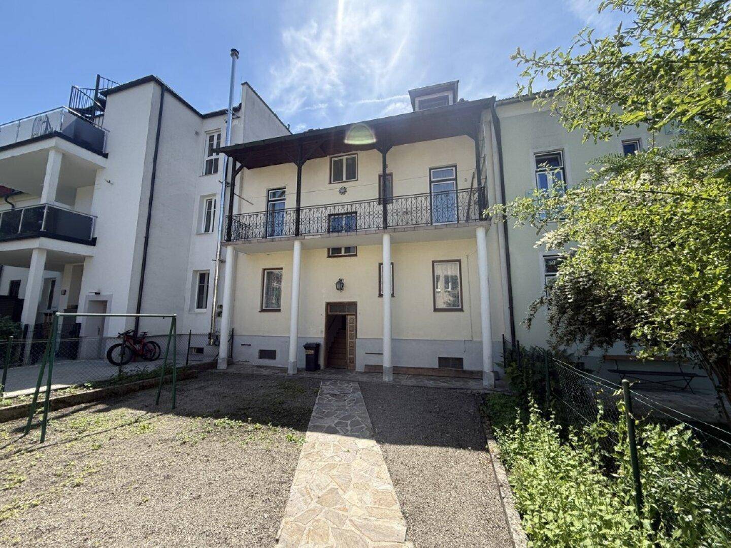 Rückseite eines Gebäudes mit großem Balkon, Säulen und einem kleinen Gartenbereich.