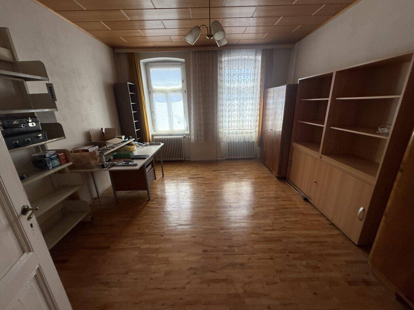Geräumiger Innenraum mit Holzparkett, Holzdecke und zwei Fenstern, eingerichtet mit Regalen und einem Schreibtisch.