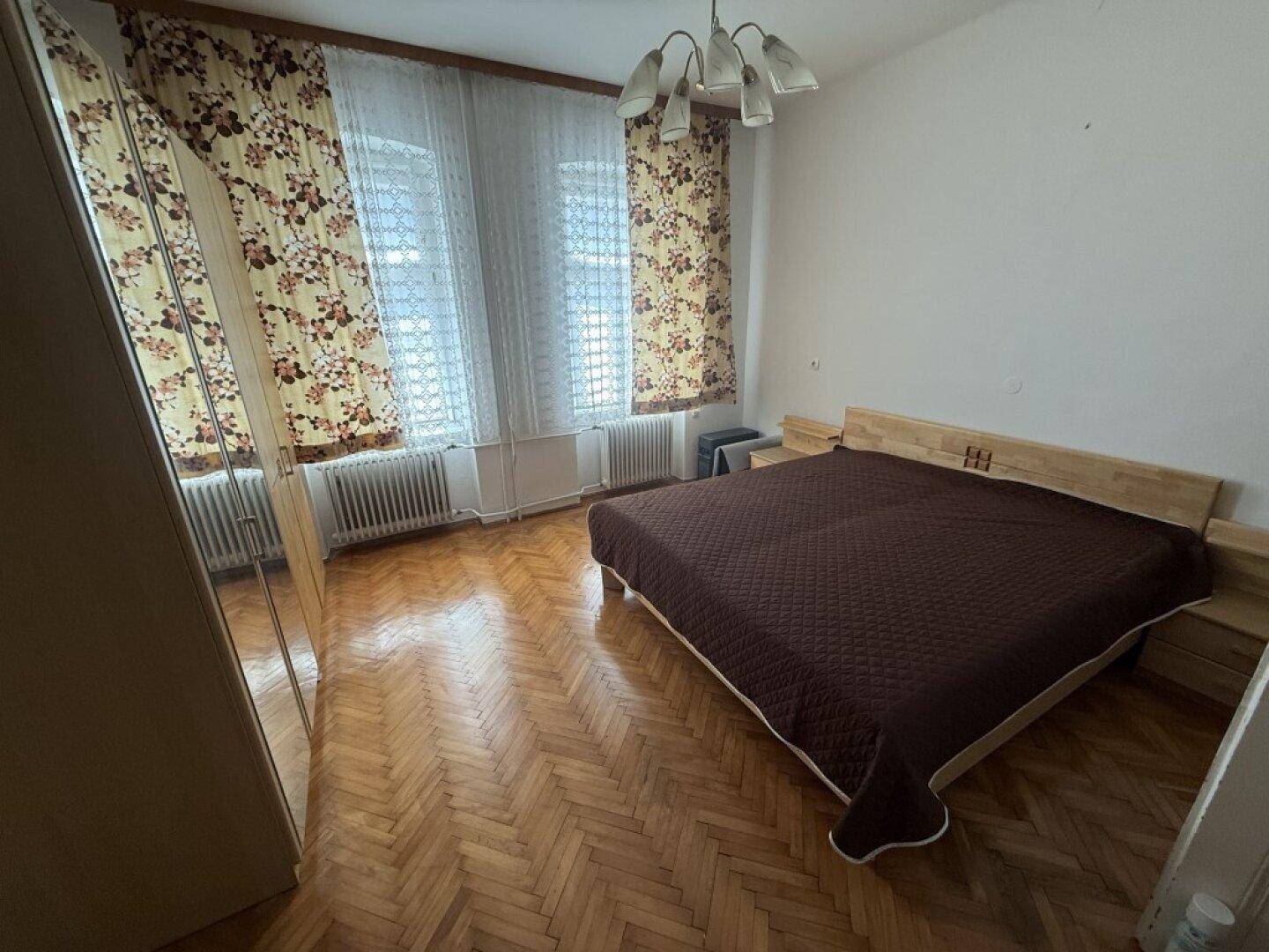 Schlafzimmer mit Holzparkett, Doppelbett und großem Spiegelschrank, beleuchtet durch zwei Fenster.