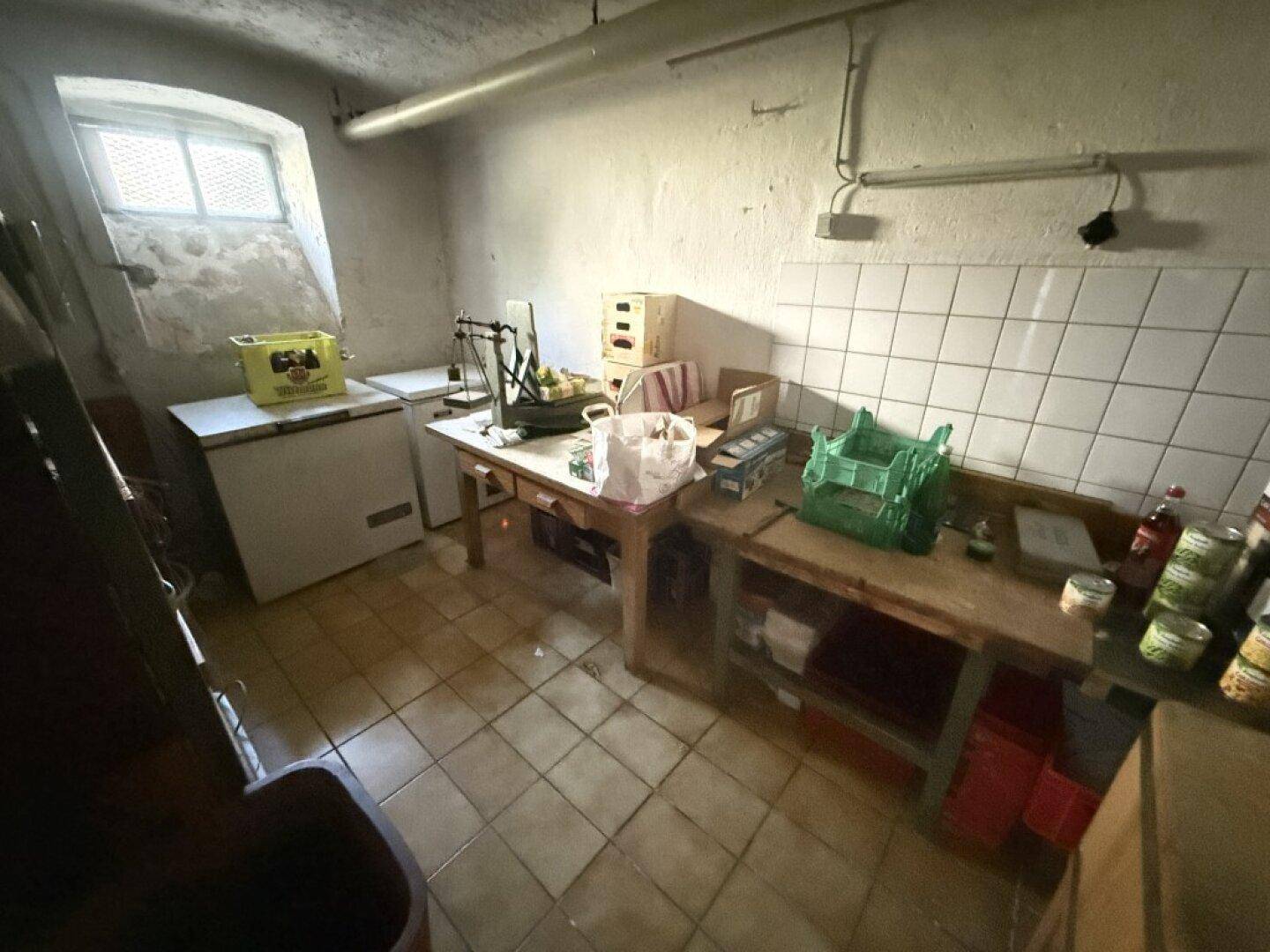 Kellerraum mit Fliesenboden, kleinen Fenstern und Arbeitsflächen, genutzt als Lager- oder Vorratsraum.