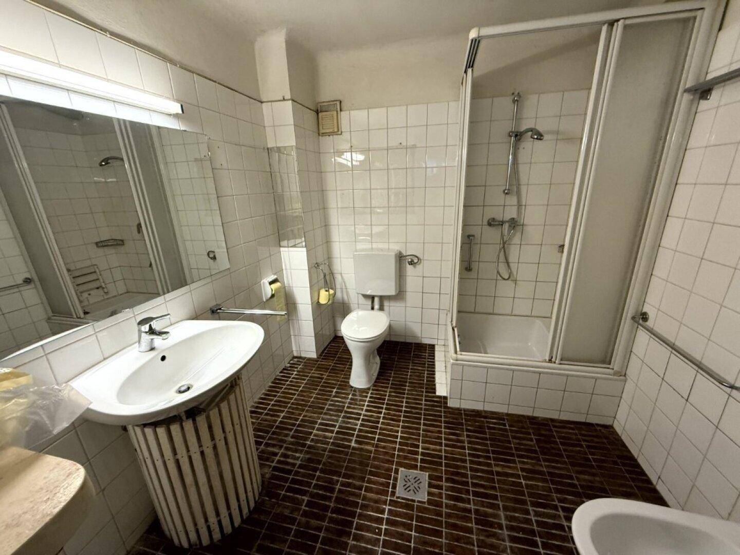 Badezimmer mit weißen Wandfliesen, dunklem Fliesenboden, Waschbecken, Toilette, Bidet und Duschkabine.