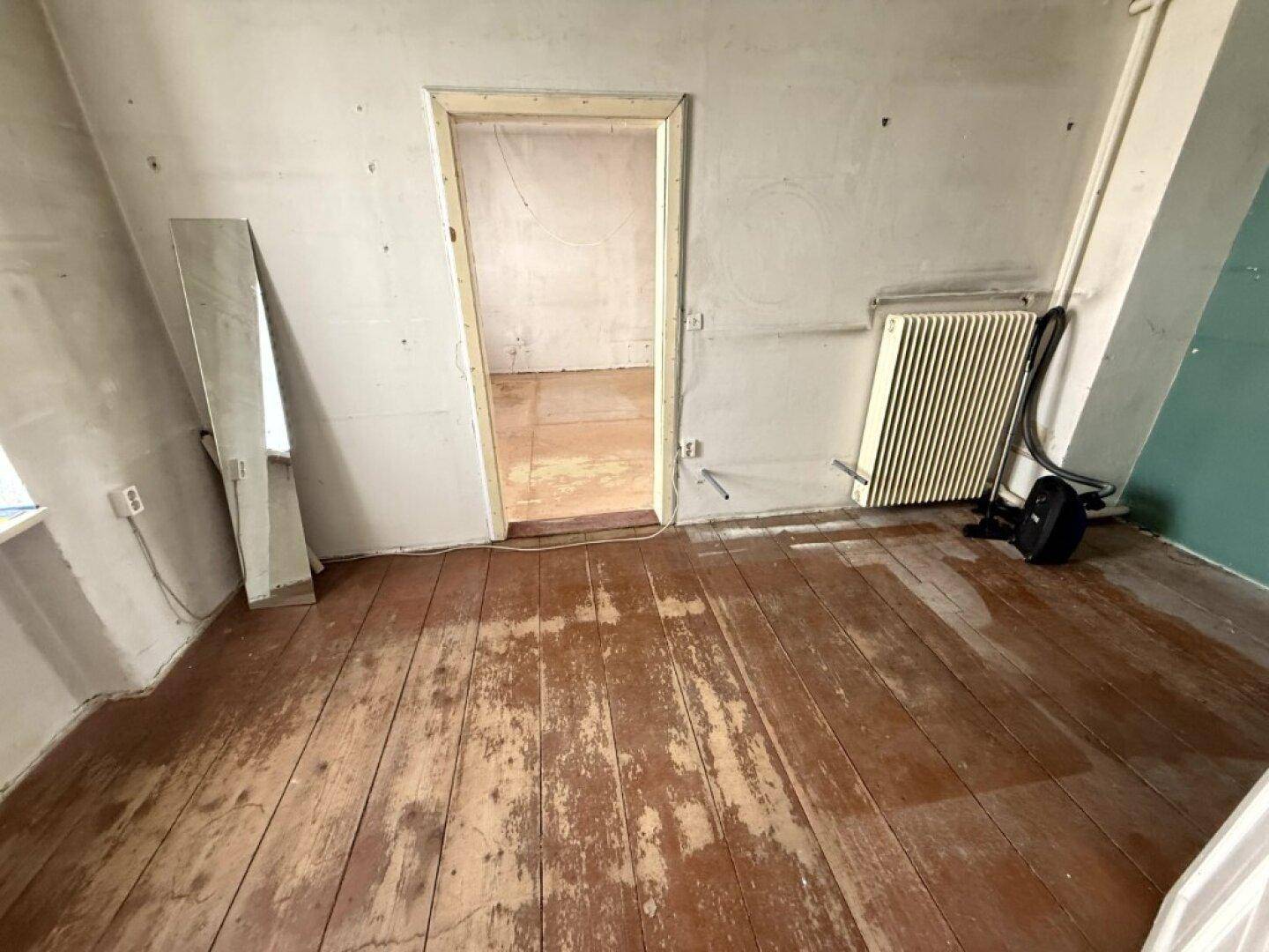 Leerer Raum mit beschädigtem Dielenboden, Heizkörper und kahlen Wänden, der Renovierungsbedarf zeigt.