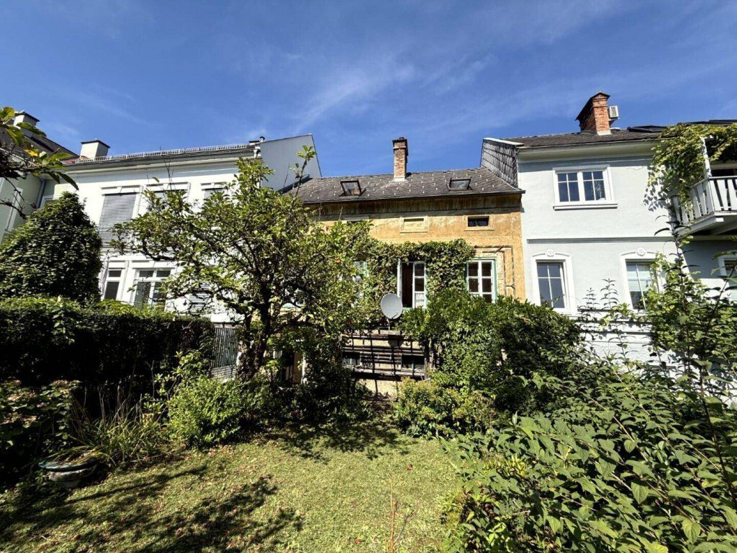 Rückseite eines Gebäudes mit gelber Fassade, umgeben von einem grünen Garten mit Bäumen und Sträuchern.