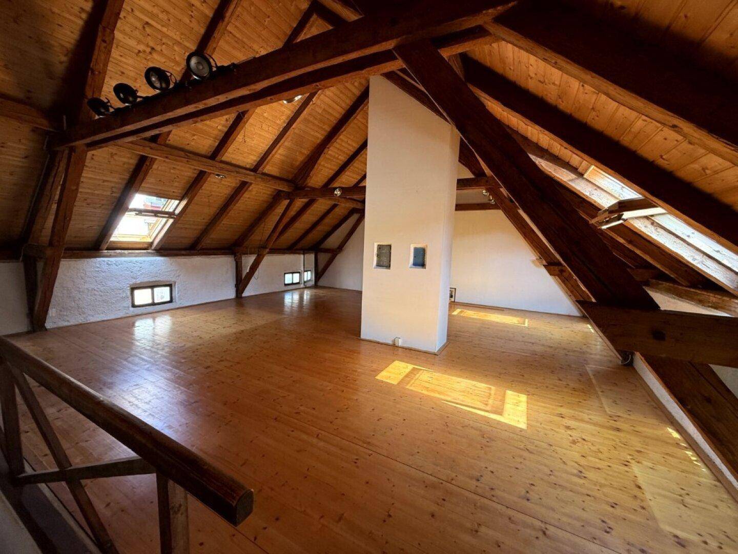 Geräumiger Dachbodenraum mit sichtbaren Holzbalken, Holzfußboden und Dachfenstern, der viel Licht bietet.
