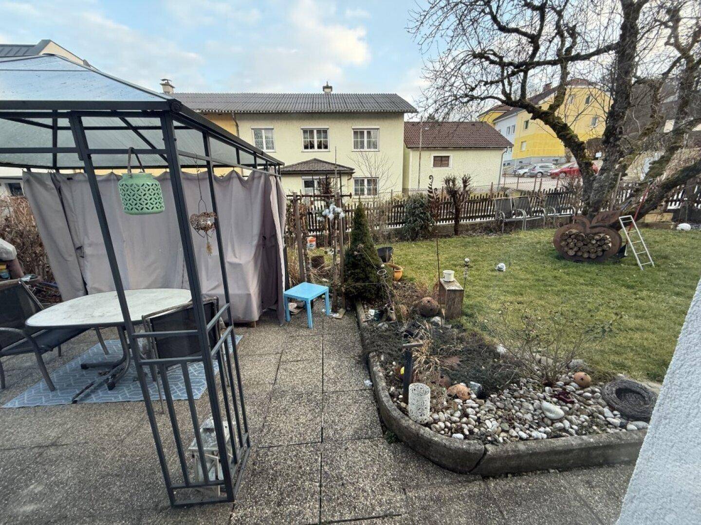 Gepflegter Garten mit gepflasterter Terrasse, Gartenlaube und Baumbestand.
