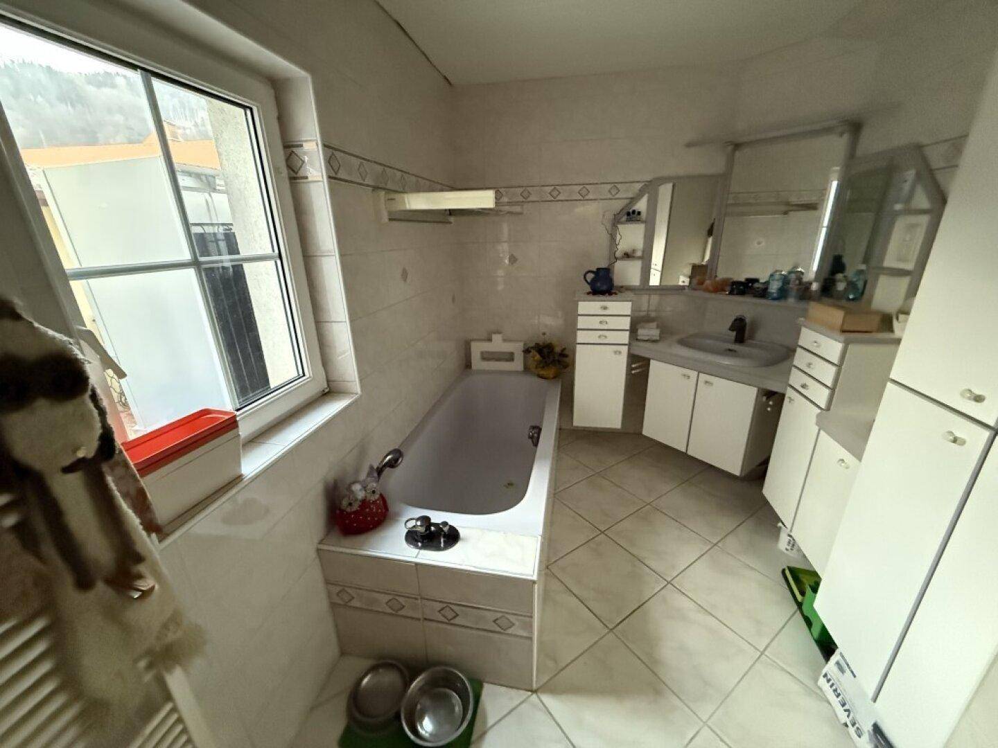 Helles Badezimmer mit Badewanne, Waschbecken und Fenster, ausgestattet mit weißen Fliesen.