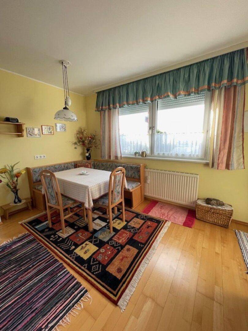 Gemütlicher Essbereich mit Eckbank, Holztisch und farbenfrohem Teppich vor dem Fenster.