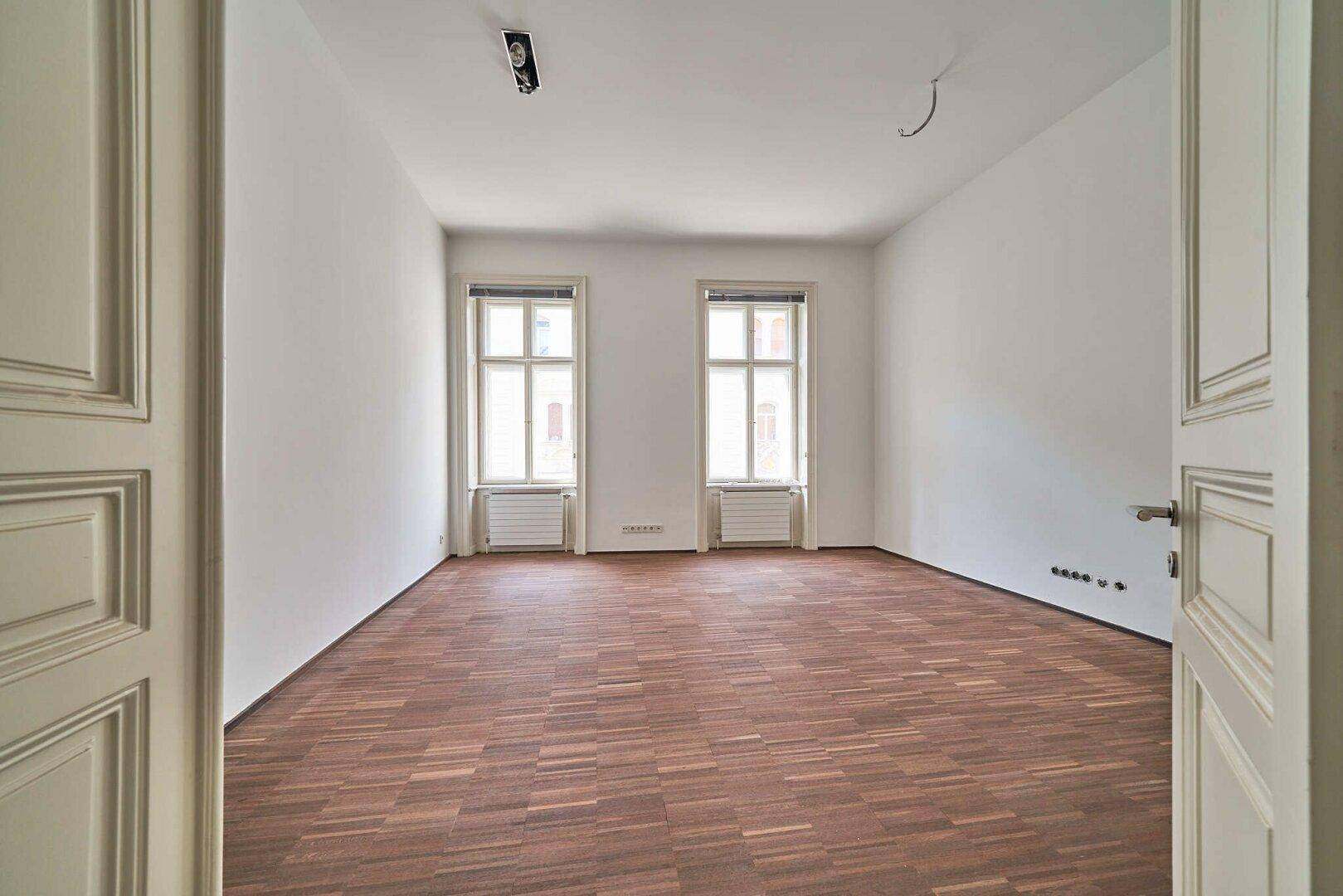 Heller, leerer Raum mit Holzboden und zwei Fenstern, bereit für die individuelle Einrichtung.