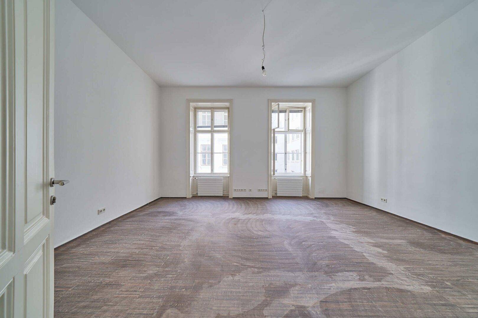 Leerer Raum mit zwei hohen Fenstern und Holzboden, der auf die Fertigstellung wartet.
