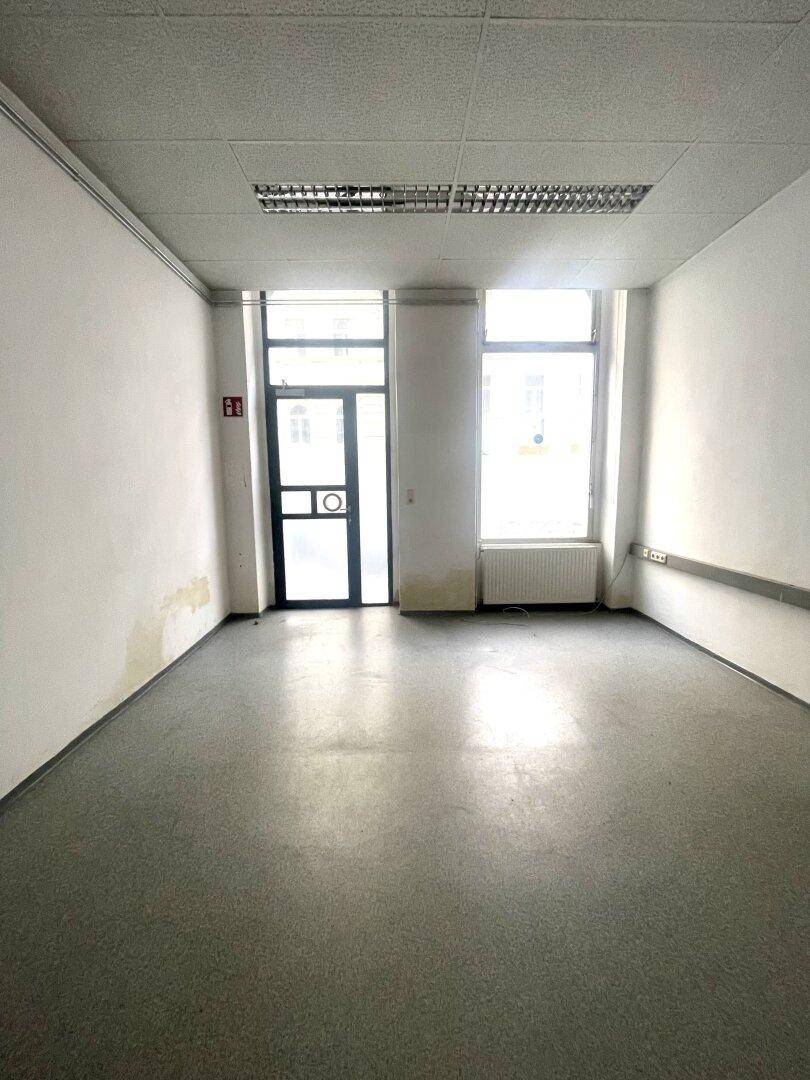 Leerer Raum mit großen Fenstern, einem Heizkörper und einer abgehängten Decke mit Beleuchtung.