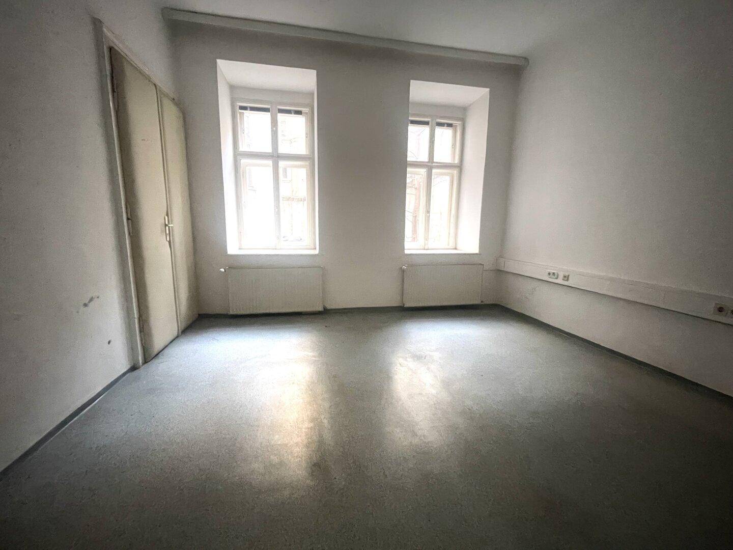 Heller, leerer Raum mit zwei Fenstern, Heizkörpern und einem einfachen Bodenbelag, bereit zur Nutzung.