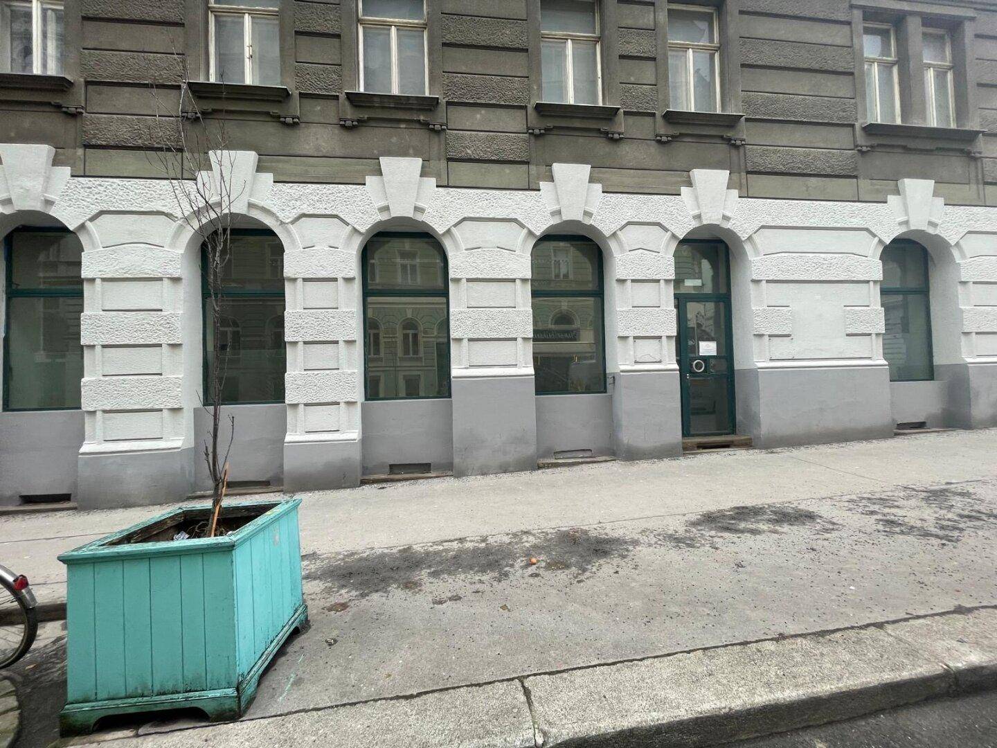 Nahaufnahme der Fassade eines Gebäudes im Erdgeschoss mit großen Bogenfenstern und einem Pflanzenkübel.