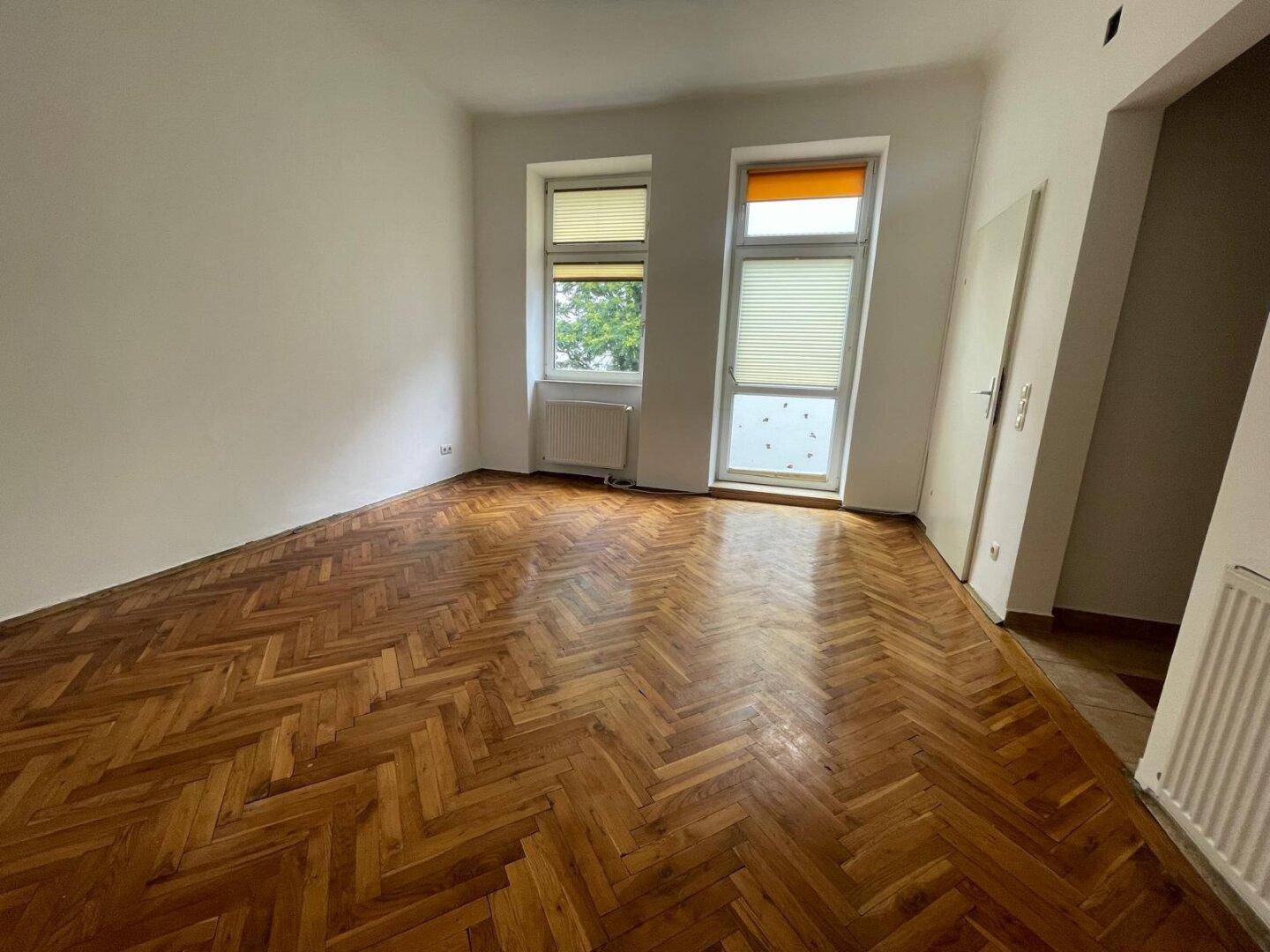 Geräumiges Zimmer mit klassischem Fischgrätparkett und zwei Fenstern mit Jalousien.