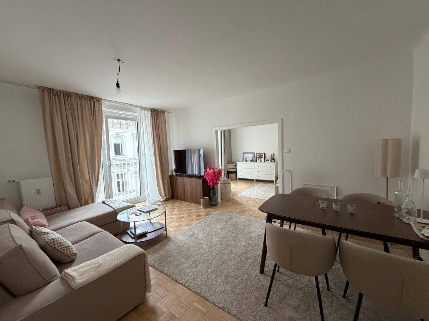 Heller Wohn-Essbereich mit Parkettboden, beigem Sofa und Esstisch, sowie Zugang zu einem französischen Balkon.